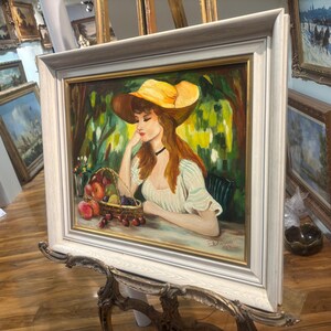 Puede incluir: Una pintura de una mujer con un sombrero amarillo y un vestido blanco, sentada en un jard&iacute;n con una cesta de fruta. La pintura est&aacute; enmarcada en un marco blanco.