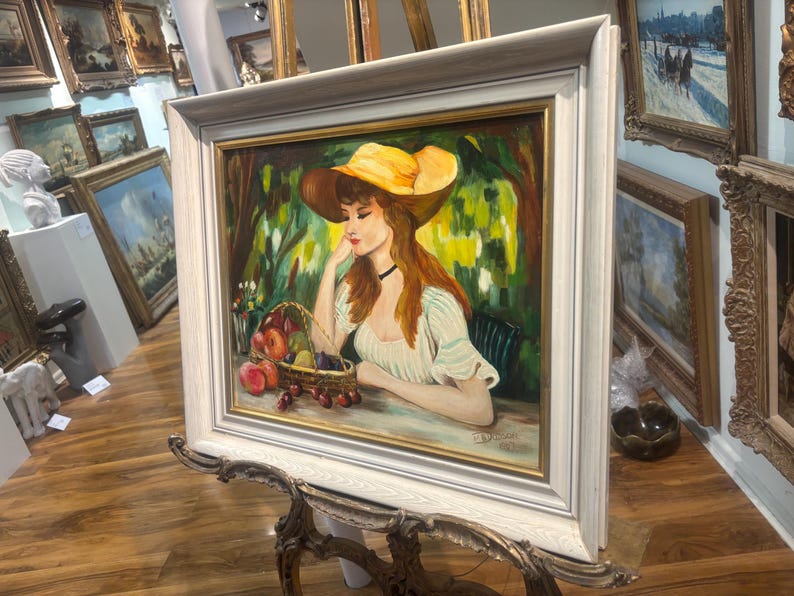 Puede incluir: Una pintura de una mujer con un sombrero amarillo y un vestido blanco, sentada en un jard&iacute;n con una cesta de fruta. La pintura est&aacute; enmarcada en un marco blanco.