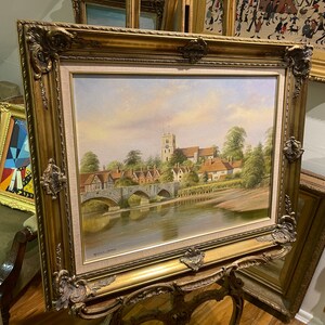 May include: A framed oil painting depicting a picturesque English village scene with a bridge over a river and a church in the background. The painting is in a gold ornate frame.