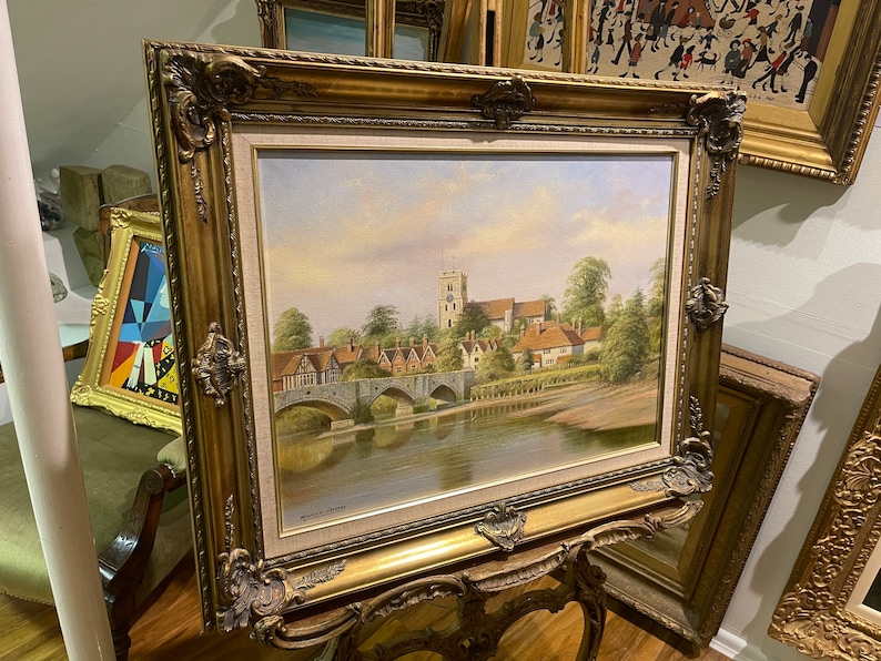 May include: A framed oil painting depicting a picturesque English village scene with a bridge over a river and a church in the background. The painting is in a gold ornate frame.