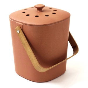 May include: A terracotta-colored compost bin with a lid featuring ventilation holes. It has a wooden handle for carrying. The bin is designed for kitchen or garden use, promoting sustainable waste management.