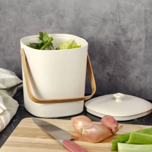 May include: A cream-colored compost bin with a speckled texture and a light brown wooden handle. The bin has a matching lid placed beside it. Inside the bin are green celery stalks. A chef's knife and two shallots sit on a wooden cutting board.