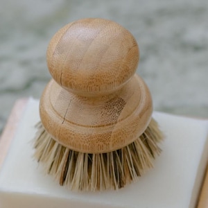 May include: A natural bamboo dish brush with a round handle and a base of bristles. The brush is resting on a white bar of soap. The bamboo has a light brown color and visible wood grain.