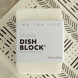May include: A rectangular, off-white Vegan Dish Block solid dishwashing soap bar with a white label. The label reads "NO TOX LIFE" and "VEGAN DISH BLOCK" with the weight of 5.9 oz and 167 g. The soap rests on a patterned surface.