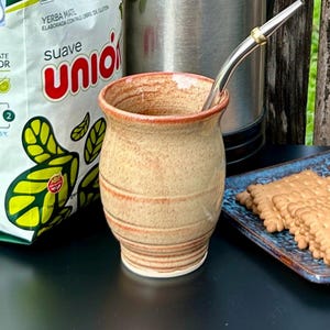 May include: A beige ceramic mate gourd with a speckled texture and horizontal ridges, featuring a reddish-brown rim and base. A silver-colored metal bombilla straw with a gold-toned ring is inserted into the top of the gourd.