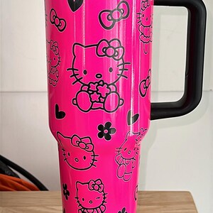May include: A pink Hello Kitty travel mug with a black handle and lid. The mug has a black and white Hello Kitty design with bows and hearts. The mug is on a silver base with a black cord.