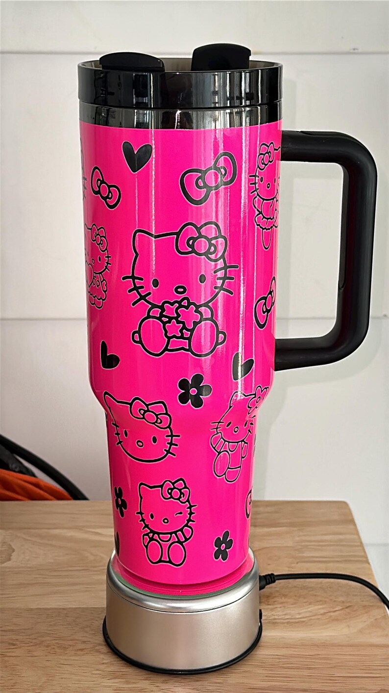 May include: A pink Hello Kitty travel mug with a black handle and lid. The mug has a black and white Hello Kitty design with bows and hearts. The mug is on a silver base with a black cord.