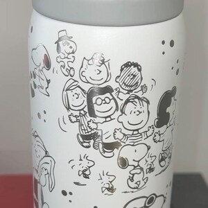 May include: White water bottle featuring a gray lid and handle. The bottle is decorated with black and gold line drawings of Peanuts characters. The design includes Snoopy, Charlie Brown, and other characters.