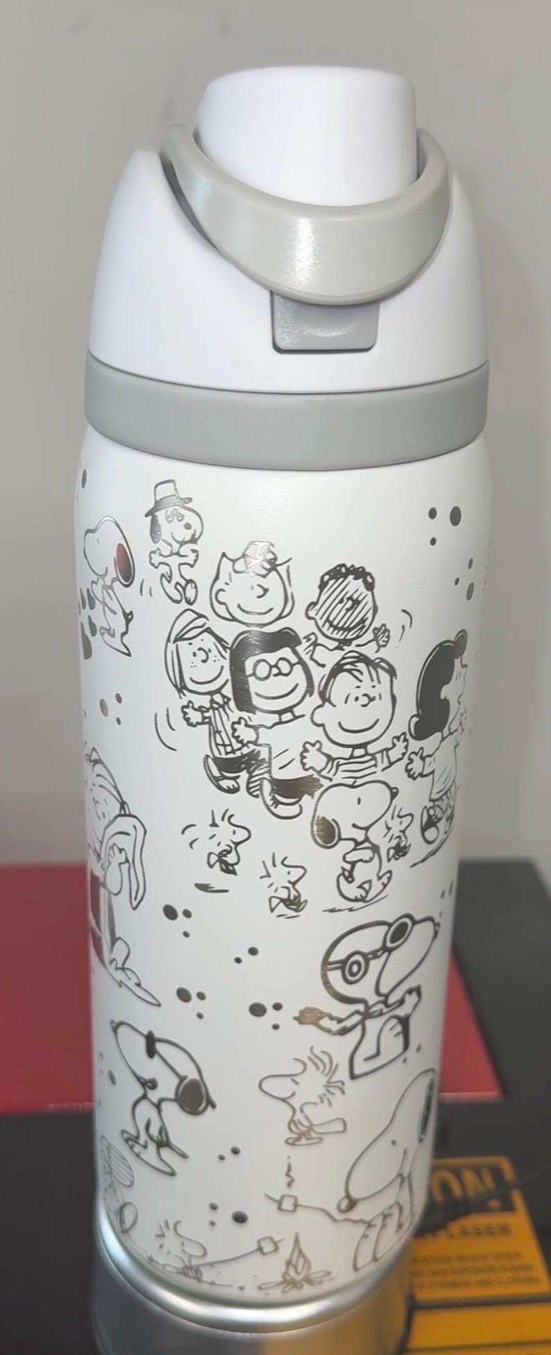 May include: White water bottle featuring a gray lid and handle. The bottle is decorated with black and gold line drawings of Peanuts characters. The design includes Snoopy, Charlie Brown, and other characters.