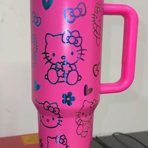 May include: Pink stainless steel travel mug with a black Hello Kitty design. The mug has a handle and a lid.