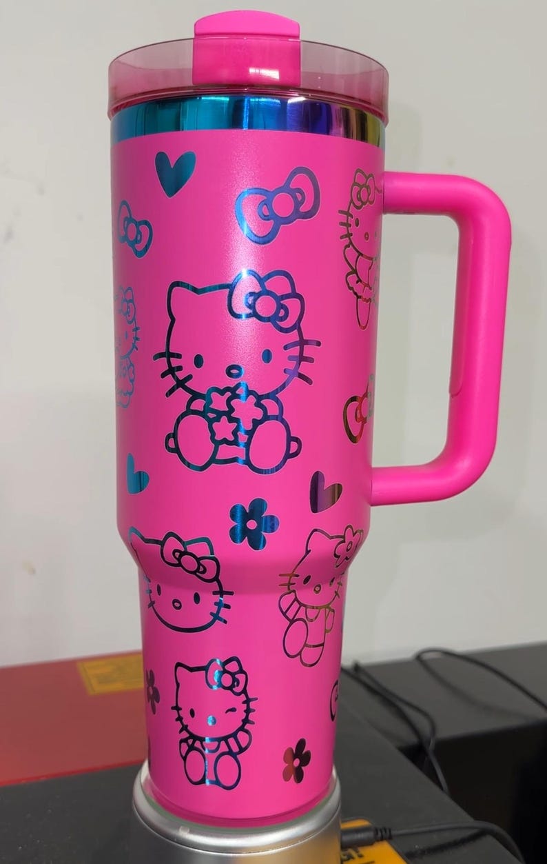 May include: Pink stainless steel travel mug with a black Hello Kitty design. The mug has a handle and a lid.