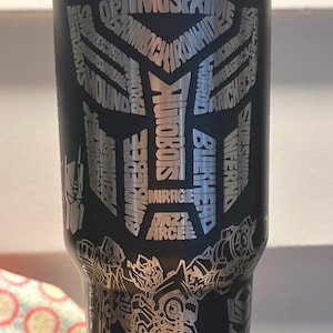 May include: Black stainless steel tumbler with a silver graphic of a robot and the words "TRANSFORMERS" and names of characters from the Transformers franchise.