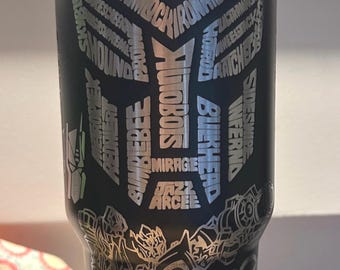 May include: Black stainless steel tumbler with a silver graphic of a robot and the words "TRANSFORMERS" and names of characters from the Transformers franchise.