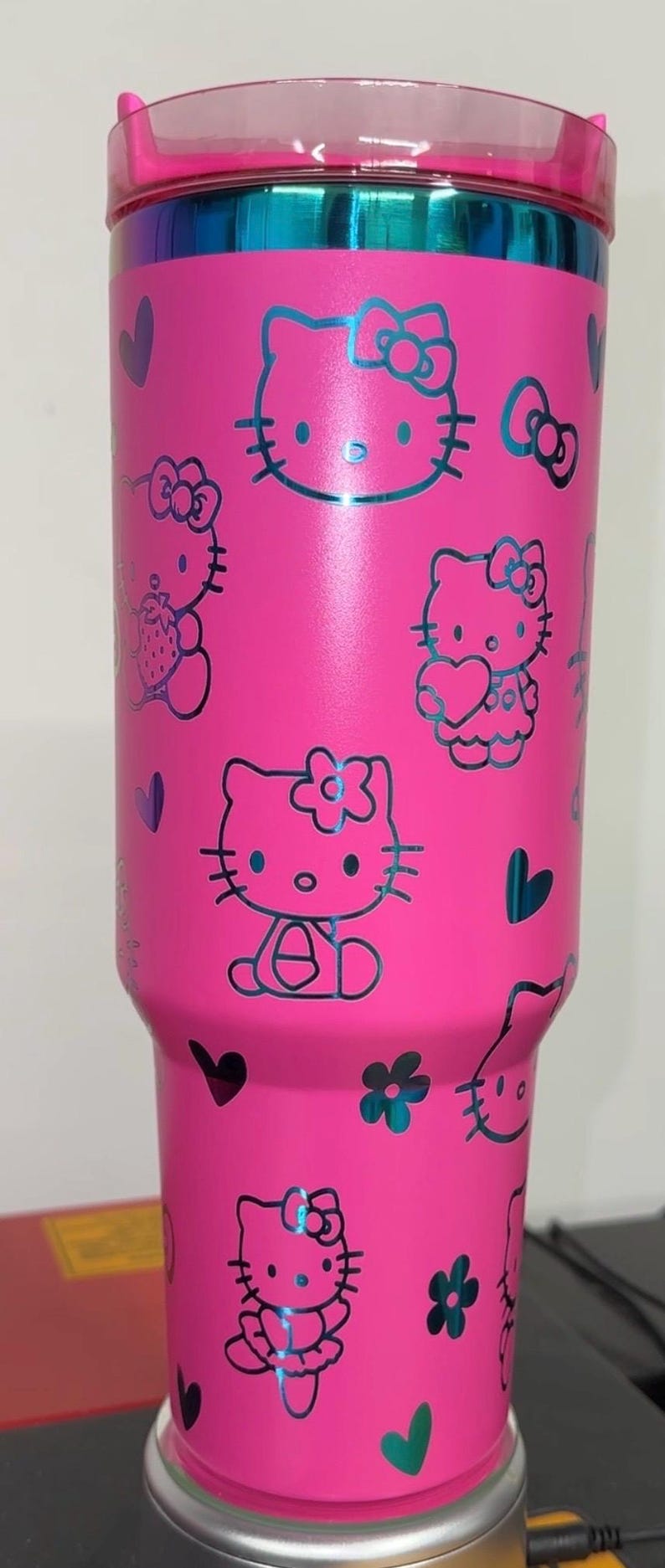 May include: Pink stainless steel tumbler with a black Hello Kitty design. The tumbler features multiple Hello Kitty characters with hearts and flowers.