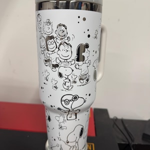 May include: A white travel mug featuring a black and white illustration of Snoopy and the Peanuts gang. The mug has a gray lid and handle, with a silver rim. The design includes various characters and scattered black dots.