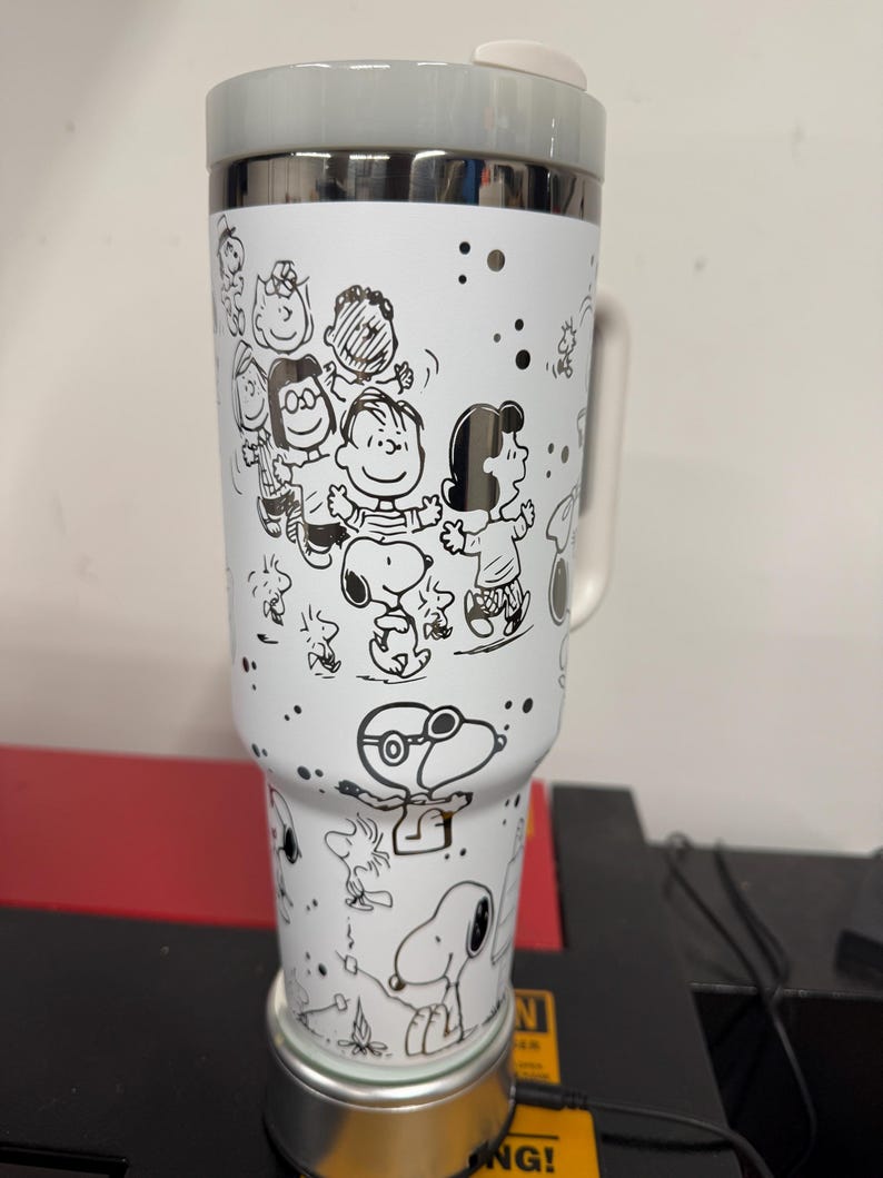 May include: A white travel mug featuring a black and white illustration of Snoopy and the Peanuts gang. The mug has a gray lid and handle, with a silver rim. The design includes various characters and scattered black dots.
