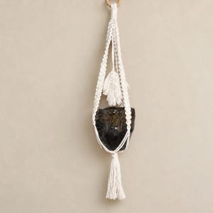 Può includere: Un portapiante in macramè realizzato in corda di cotone color bianco sporco con intricati motivi a nodo, una nappa decorativa centrale e un vaso in ceramica scuro, lucido e maculato sospeso nella struttura intrecciata.