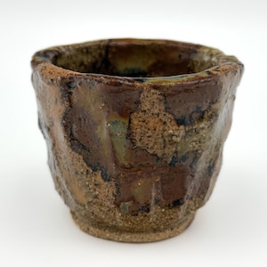 Può includere: Una piccola tazza in ceramica modellata a mano con una superficie irregolare e strutturata. L'esterno presenta uno smalto maculato nei toni del marrone scuro, marrone chiaro e verde spento, con una base ruvida e sabbiosa.