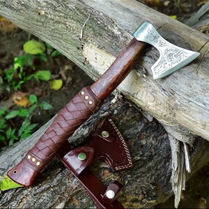 Puede incluir: Un hacha artesanal con mango de madera y hoja plateada con intrincados grabados. El mango est&aacute; envuelto en cuero marr&oacute;n con remaches de lat&oacute;n. Tambi&eacute;n se incluye una funda de cuero a juego.