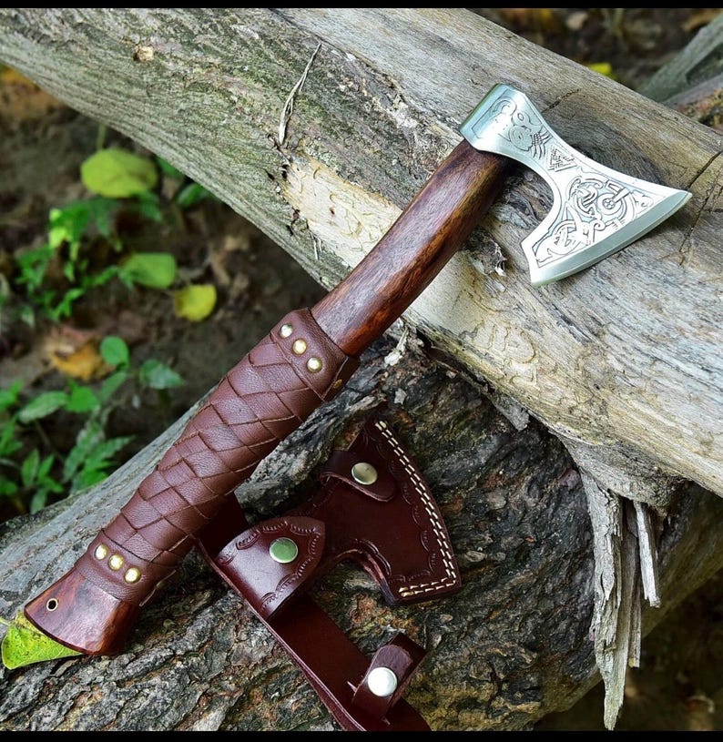 Puede incluir: Un hacha artesanal con mango de madera y hoja plateada con intrincados grabados. El mango est&aacute; envuelto en cuero marr&oacute;n con remaches de lat&oacute;n. Tambi&eacute;n se incluye una funda de cuero a juego.