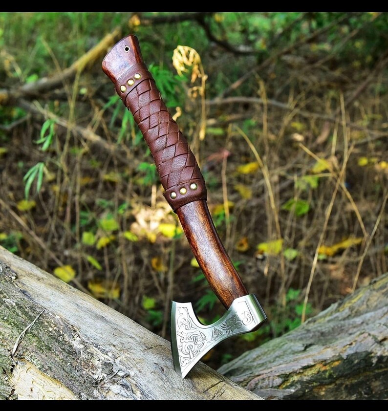 Puede incluir: Un hacha artesanal con mango de madera y empu&ntilde;adura de cuero marr&oacute;n. La cabeza del hacha es plateada con un dise&ntilde;o grabado. El hacha est&aacute; apoyada en un tronco en un entorno natural.