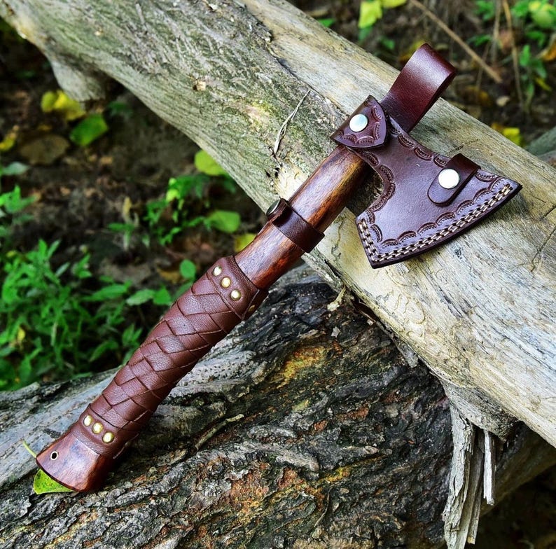 Puede incluir: Un hacha artesanal con un mango de madera marr&oacute;n oscuro y un agarre de cuero trenzado. La cabeza del hacha est&aacute; cubierta con una funda de cuero marr&oacute;n. El hacha descansa sobre un tronco desgastado, mostrando su dise&ntilde;o r&uacute;stico.