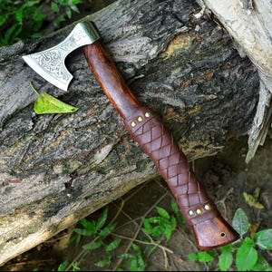 Puede incluir: Un hacha artesanal con una hoja plateada pulida con un dise&ntilde;o grabado. El mango es de madera oscura y est&aacute; envuelto en cuero marr&oacute;n. Remaches de lat&oacute;n aseguran el agarre de cuero. El hacha descansa sobre un tronco desgastado.