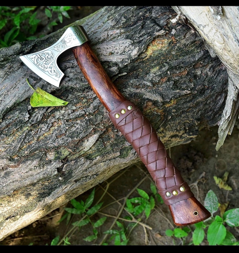 Puede incluir: Un hacha artesanal con una hoja plateada pulida con un dise&ntilde;o grabado. El mango es de madera oscura y est&aacute; envuelto en cuero marr&oacute;n. Remaches de lat&oacute;n aseguran el agarre de cuero. El hacha descansa sobre un tronco desgastado.