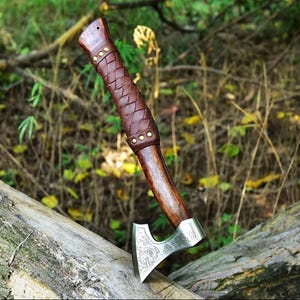 Puede incluir: Un hacha artesanal con mango de madera y empu&ntilde;adura de cuero marr&oacute;n. La cabeza del hacha es plateada con un dise&ntilde;o grabado. El hacha est&aacute; apoyada sobre un tronco en un entorno natural al aire libre.