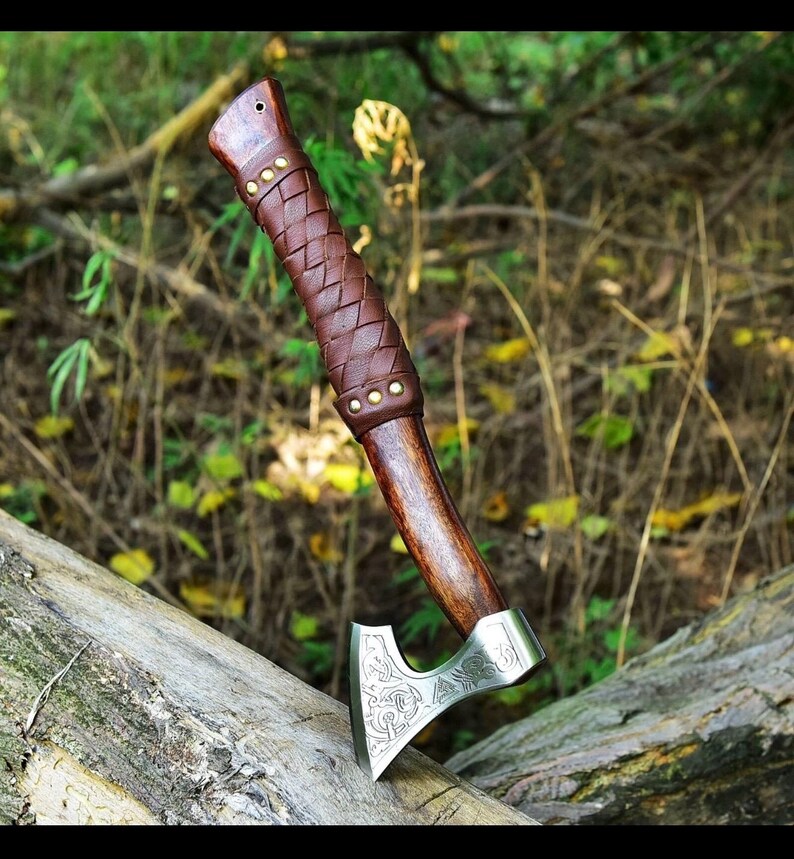 Puede incluir: Un hacha artesanal con mango de madera y empu&ntilde;adura de cuero marr&oacute;n. La cabeza del hacha es plateada con un dise&ntilde;o grabado. El hacha est&aacute; apoyada sobre un tronco en un entorno natural al aire libre.