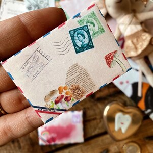 May include: A vintage-style envelope with a red, white, and blue border. The envelope has a postage stamp with a portrait of a woman and the words "Via Air Mail" printed on the bottom. The envelope is decorated with a mushroom and pine cone illustration.