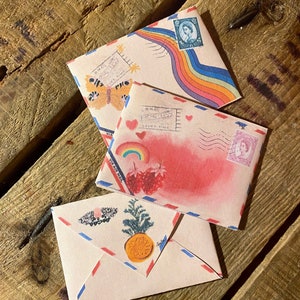 May include: Three vintage-style envelopes with colorful designs. The top envelope features a rainbow and a butterfly, the middle envelope has a pink watercolor background with strawberries and hearts, and the bottom envelope has a pineapple and a floral wreath.