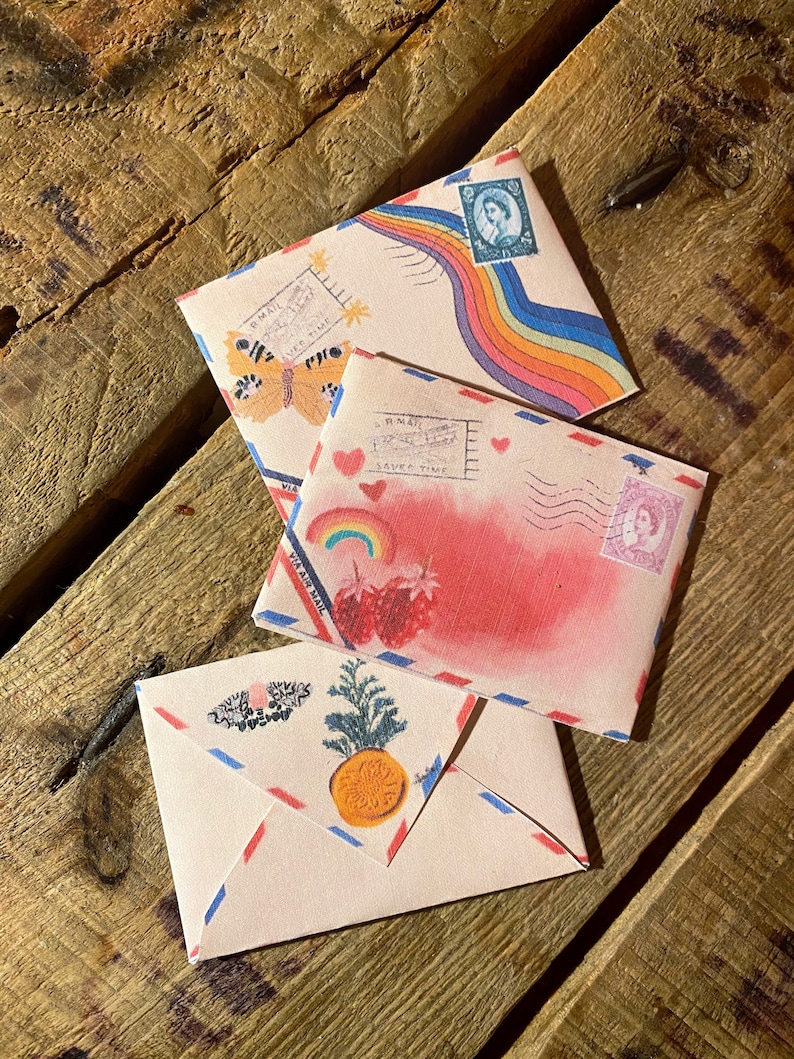 May include: Three vintage-style envelopes with colorful designs. The top envelope features a rainbow and a butterfly, the middle envelope has a pink watercolor background with strawberries and hearts, and the bottom envelope has a pineapple and a floral wreath.