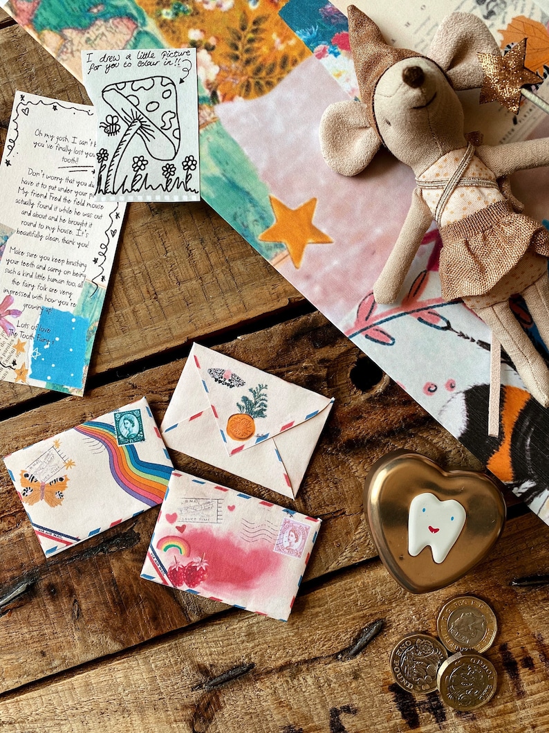 May include: A wooden table with a heart-shaped box, three coins, and three envelopes decorated with rainbows, butterflies, and strawberries. The envelopes are addressed to a tooth fairy. A handwritten note with a drawing of a mushroom is also on the table.