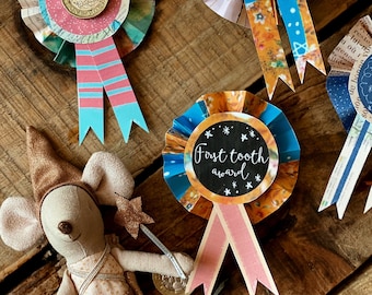 May include: A collection of handmade award ribbons with colorful paper rosettes and decorative ribbons. One ribbon features a coin, while others have text that reads "Super shiny tooth Award" and "First tooth award". A small mouse doll is also present.