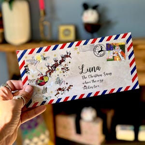 Puede incluir: Un sobre rectangular con borde de rayas rojas y azules que presenta un gráfico de Santa en un trineo tirado por renos, mariposas y purpurina dorada. Está dirigido a Luna, The Christmas Snug where the cozy teddies live.