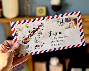 May include: A rectangular envelope with a red and blue striped border featuring a graphic of Santa in a sleigh pulled by reindeer, butterflies, and gold glitter. It is addressed to Luna at The Christmas Snug where the cozy teddies live.