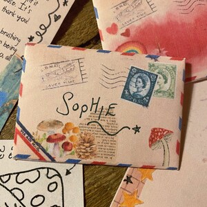 May include: A vintage-style envelope with a red, white, and blue border. The envelope has a stamp that says "IR MAIL SAVES TIME" and a stamp with a portrait of a woman. The envelope is addressed to "Sophie" and has a small star drawn on it.