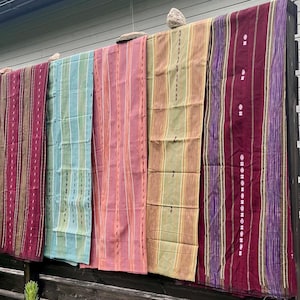 以下が含まれることがあります： 木製のフェンスに掛けられた、5つのカラフルな縦縞模様の織物。赤、緑、ピンク、紫など、さまざまな色があります。これらの生地は、装飾品や工芸品として使用されている可能性があります。