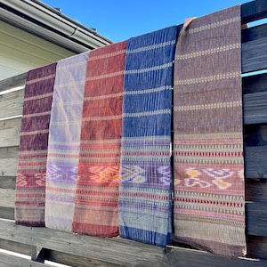 以下が含まれることがあります： 木製のフェンスに掛けられた、複雑な模様が施されたカラフルな織り生地のストール5枚。ストールは、紫、青、赤、茶色の色合いです。