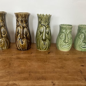 May include: Five ceramic vessels arranged in a row, each featuring a sculpted face with a nose, eyes, and a mouth. The word "Celery" is embossed near the top rim of each piece. The vessels vary in height and colour, ranging from brown to green.