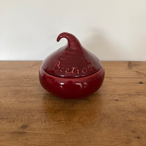 May include: A red ceramic beetroot-shaped container with a lid. The lid has a small handle and the word "Beetroot" is written on the top.