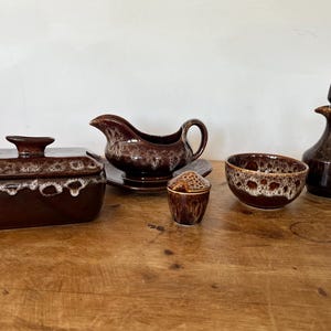 May include: A collection of brown ceramic tableware featuring a rectangular butter dish with a lid, a gravy boat on a matching saucer, a small lidded jar, a round bowl, and a cruet with a handle, all finished with a white drip glaze pattern.