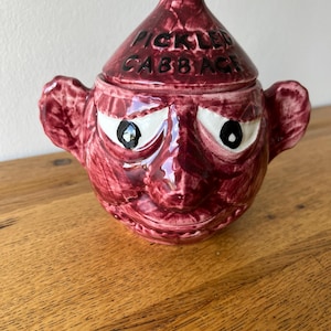 May include: A dark red ceramic jar with a lid, shaped like a face with prominent ears. The lid features the text PICKLED CABBAGE in black letters. The jar has white eyes with black pupils and a textured, glossy finish throughout the entire piece.