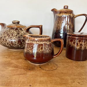 May include: A ceramic tea set featuring a teapot, a tall coffee pot, a creamer, and a lidded sugar bowl. Each piece displays a two-tone glaze with a dark brown base and a mottled, cream-coloured drip pattern cascading from the top edges.