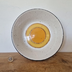 May include: A round ceramic bowl with a speckled off-white finish and a dark rim. The centre features a circular, glossy yellow design. A small silver coin is placed to the left of the bowl for scale, showing the relative size of the object.