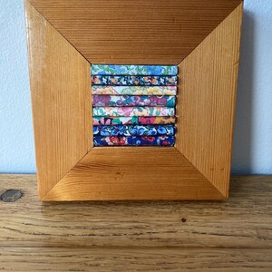 May include: A wooden frame with a square opening, displaying a colourful fabric art piece. The fabric is cut into strips and arranged in a vertical pattern, featuring floral and geometric designs in shades of blue, pink, yellow, and green.