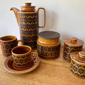May include: A vintage brown ceramic coffee set with a geometric black and gold pattern. The set includes a coffee pot, creamer, sugar bowl, two lidded jars, a cup, and a saucer. The sugar bowl has the word "SUGAR" on it.