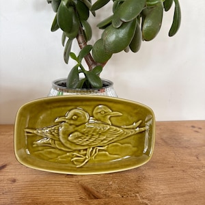 May include: An olive green ceramic rectangular tray with rounded corners featuring a raised relief design of two seagulls. The birds are depicted in profile with detailed feathers and wings, set against a smooth, glossy, monochromatic glazed surface.