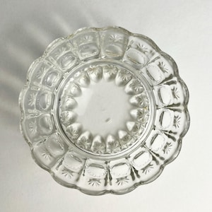 May include: A clear glass plate featuring a scalloped edge and a decorative pressed pattern. The design includes a central circular area surrounded by a ring of starburst motifs and rectangular panels with textured borders along the rim.